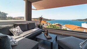 A balcony with a sofa, a coffee table, and a view of the sea and buildings. - Villa Miracle (Rogoznica)