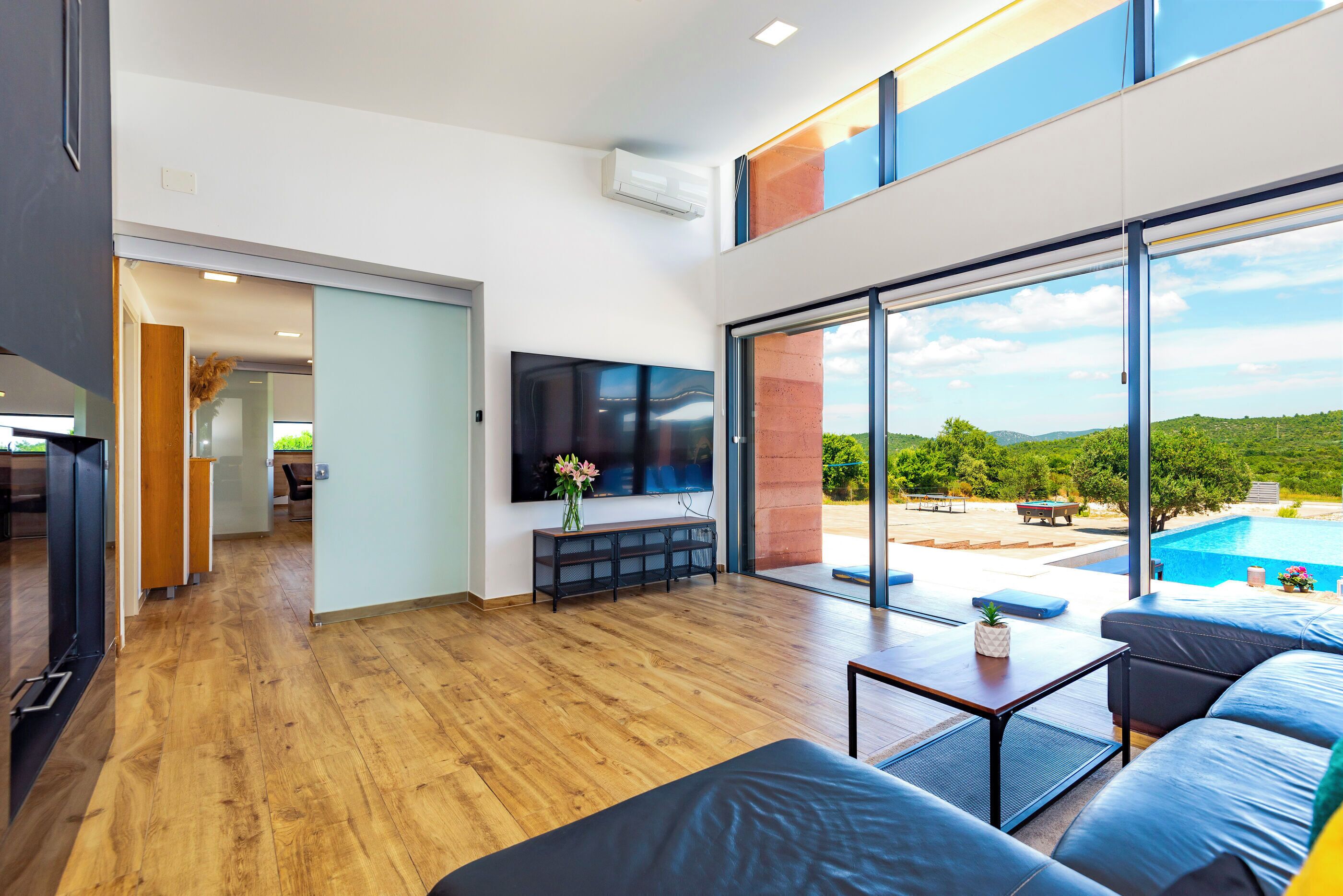 A modern living room with a large TV, wooden flooring, and a view of a swimming pool.