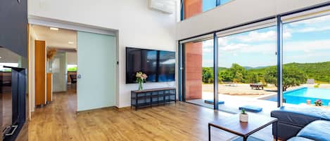 A modern living room with a large TV, wooden flooring, and a view of a swimming pool.