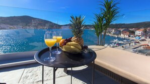 Une table avec un panier de fruits et des boissons sur un balcon donnant sur une marina et une étendue d’eau.