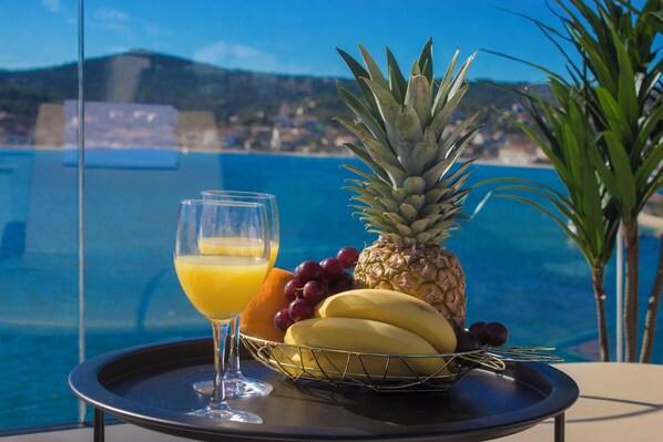 Ein Tisch mit einer Obstschale, die Bananen, Trauben und eine Ananas enthält, sowie ein Glas Orangensaft.