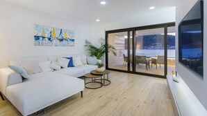 Un salon moderne avec un canapé blanc, un tableau représentant des voiliers, une plante en pot et une vue sur un espace de détente extérieur, accessible par des portes vitrées.