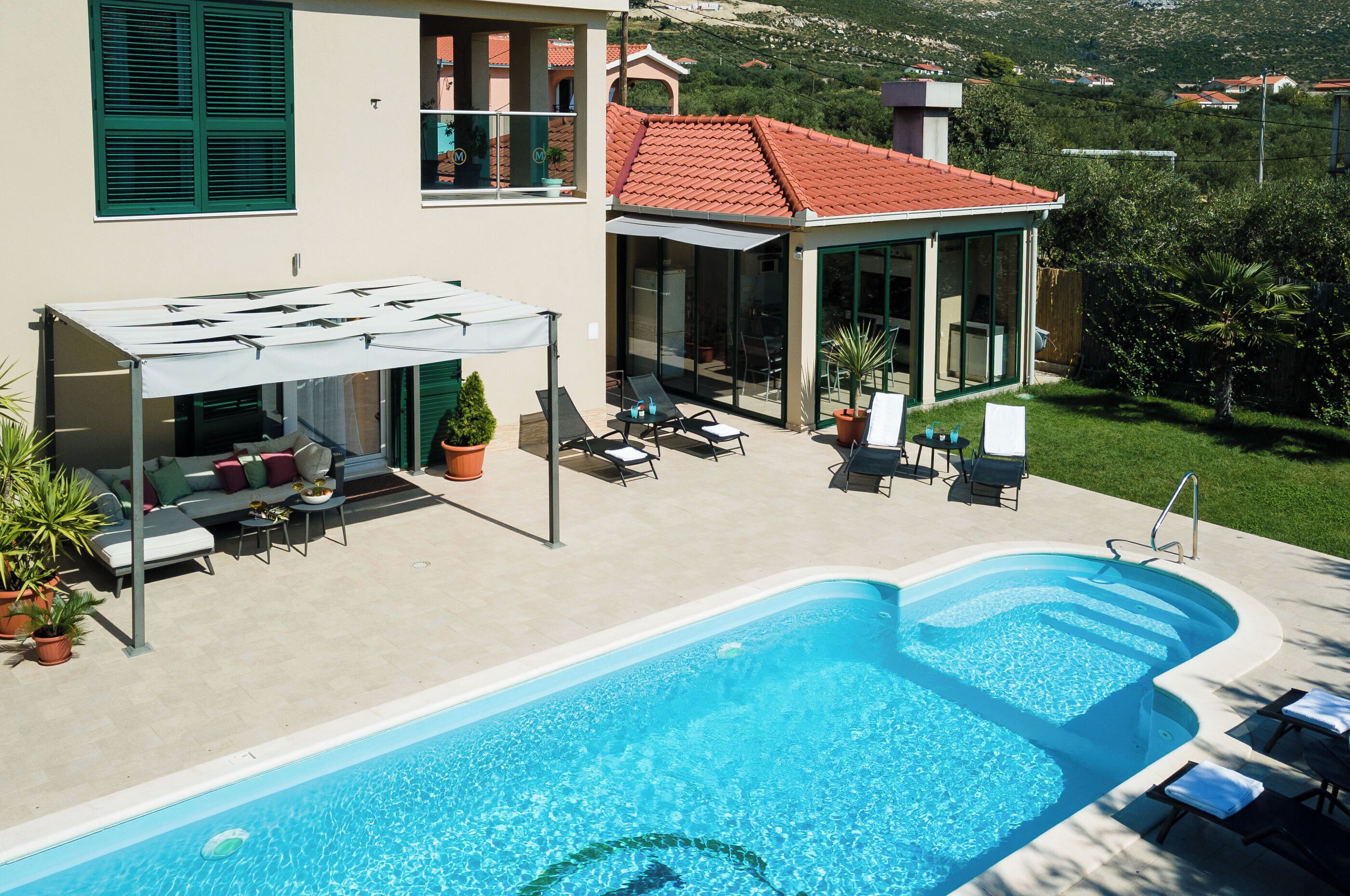 Une piscine entourée de chaises longues et d’un espace de repos couvert, le tout bordé d’une maison avec un toit de tuiles rouges et des volets verts.