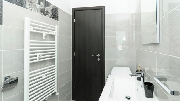 Une salle de bain moderne avec une porte en bois foncé, des murs recouverts de carreaux blancs, un lavabo blanc avec un miroir et un porte-serviettes.
