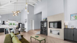 Una sala de estar moderna con un sofá verde, una mesa de centro de madera y una chimenea. En el fondo, se ve una cocina con armarios blancos y una zona de comedor.