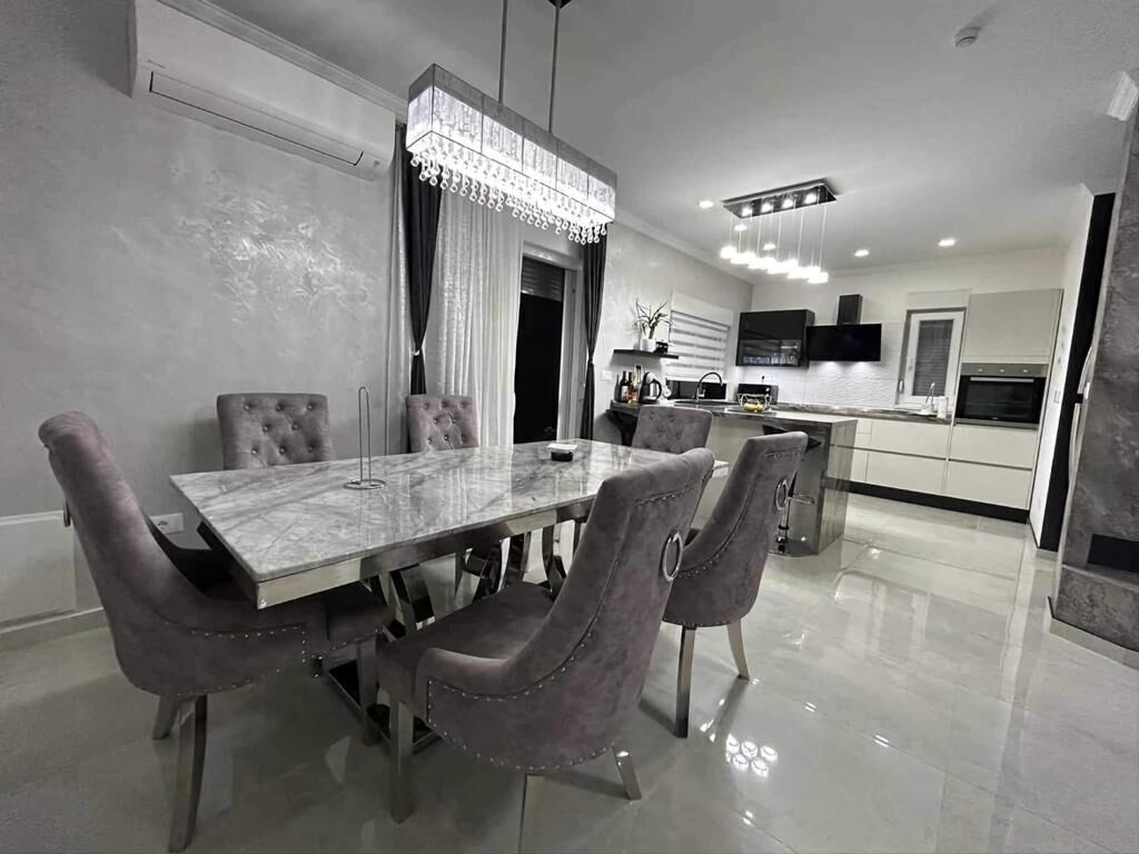 A modern dining area with a marble table and upholstered chairs, a chandelier, and a kitchen in the background.