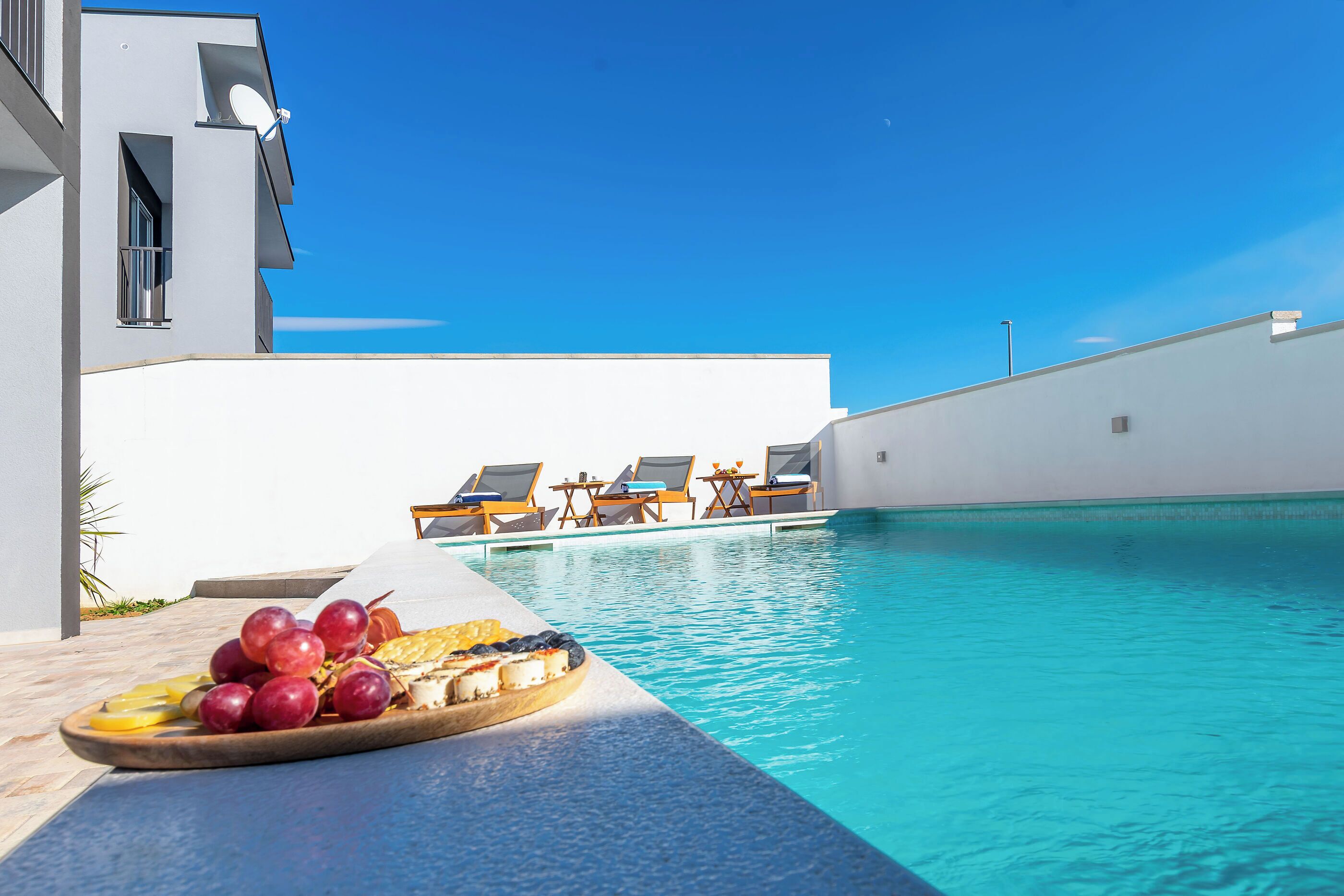 A poolside area with a tray of fruit and cheese, lounge chairs, and a modern building with a white facade.