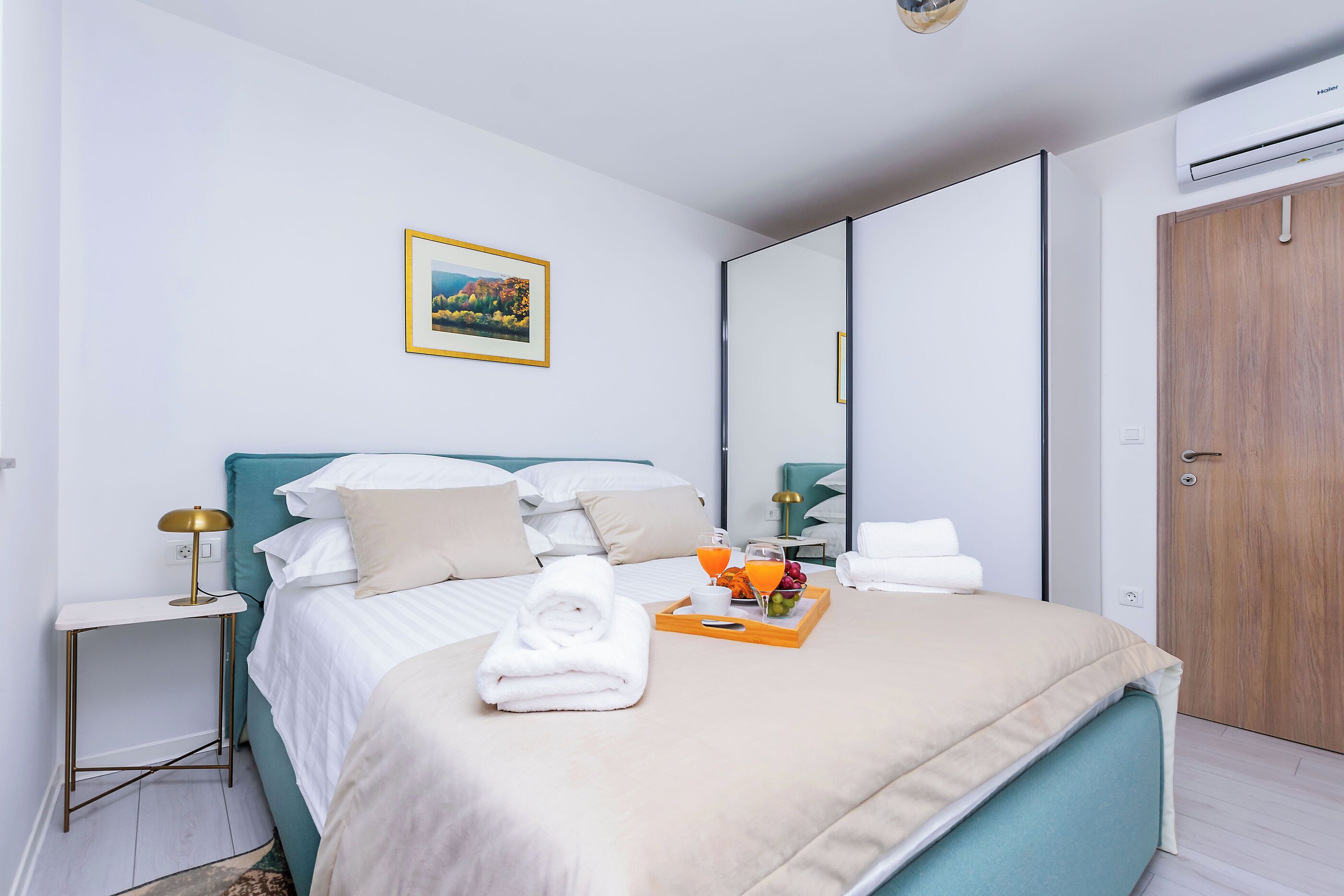 A modern hotel room with a neatly made bed, a bedside table with a lamp, a tray with fruit and juice, and a framed picture on the wall.