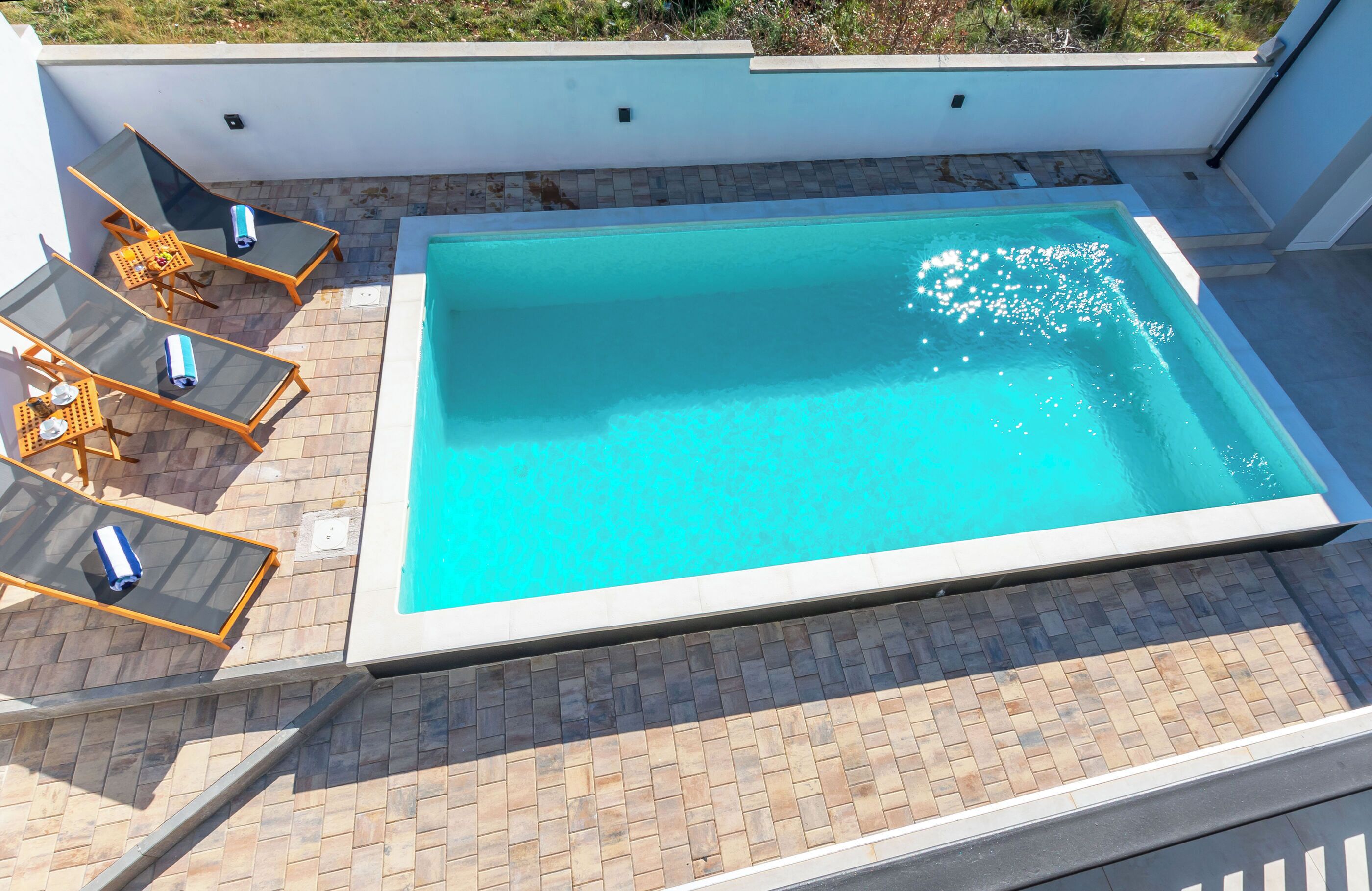 A rectangular swimming pool with a tiled deck, surrounded by lounge chairs and a glass table.