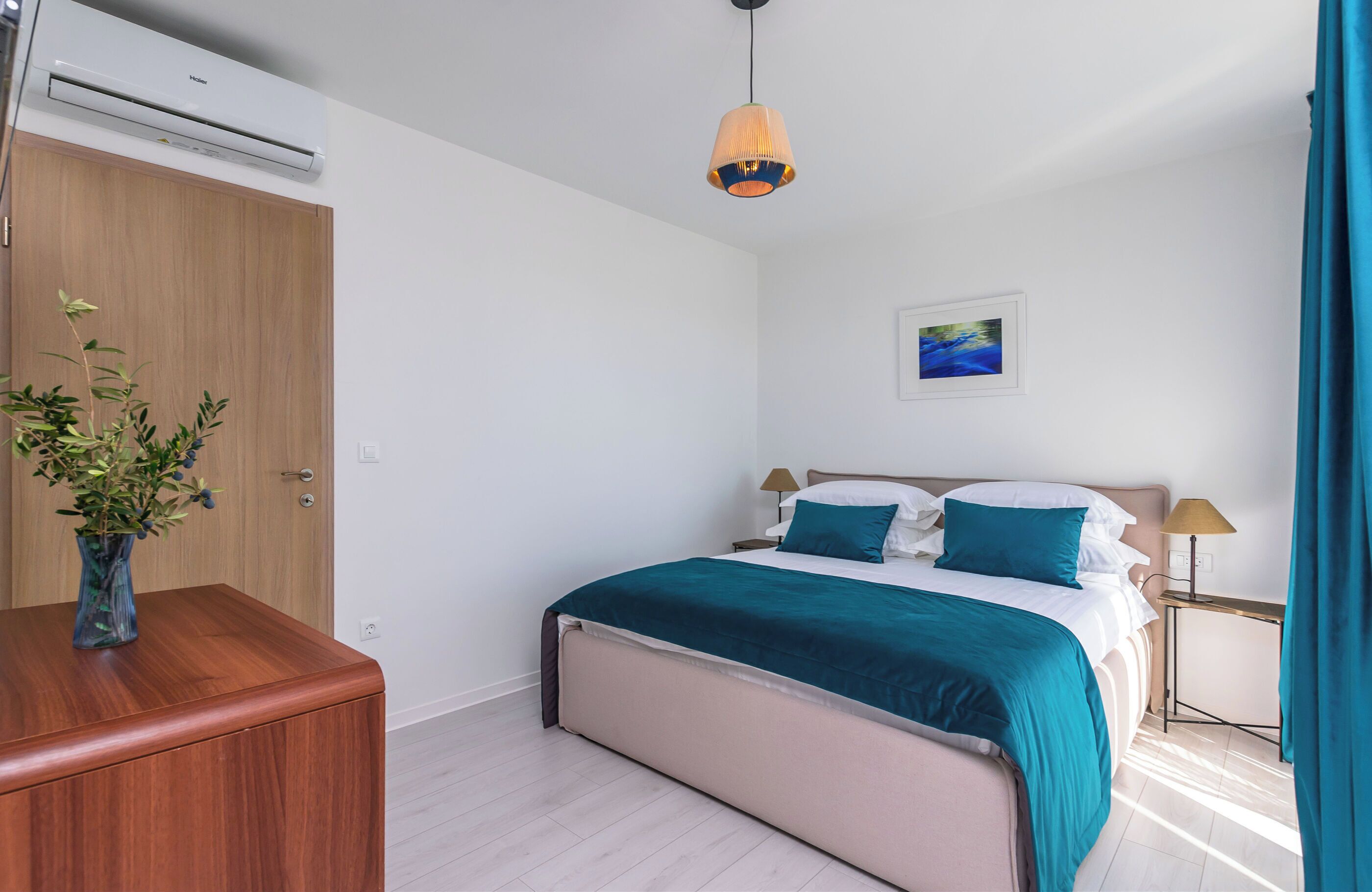 Une chambre à coucher comprenant un lit, des tables de chevet, une petite table, un climatiseur fixé au mur, une lampe suspendue, un tableau encadré et une commode en bois sur laquelle est posé un vase de fleurs.