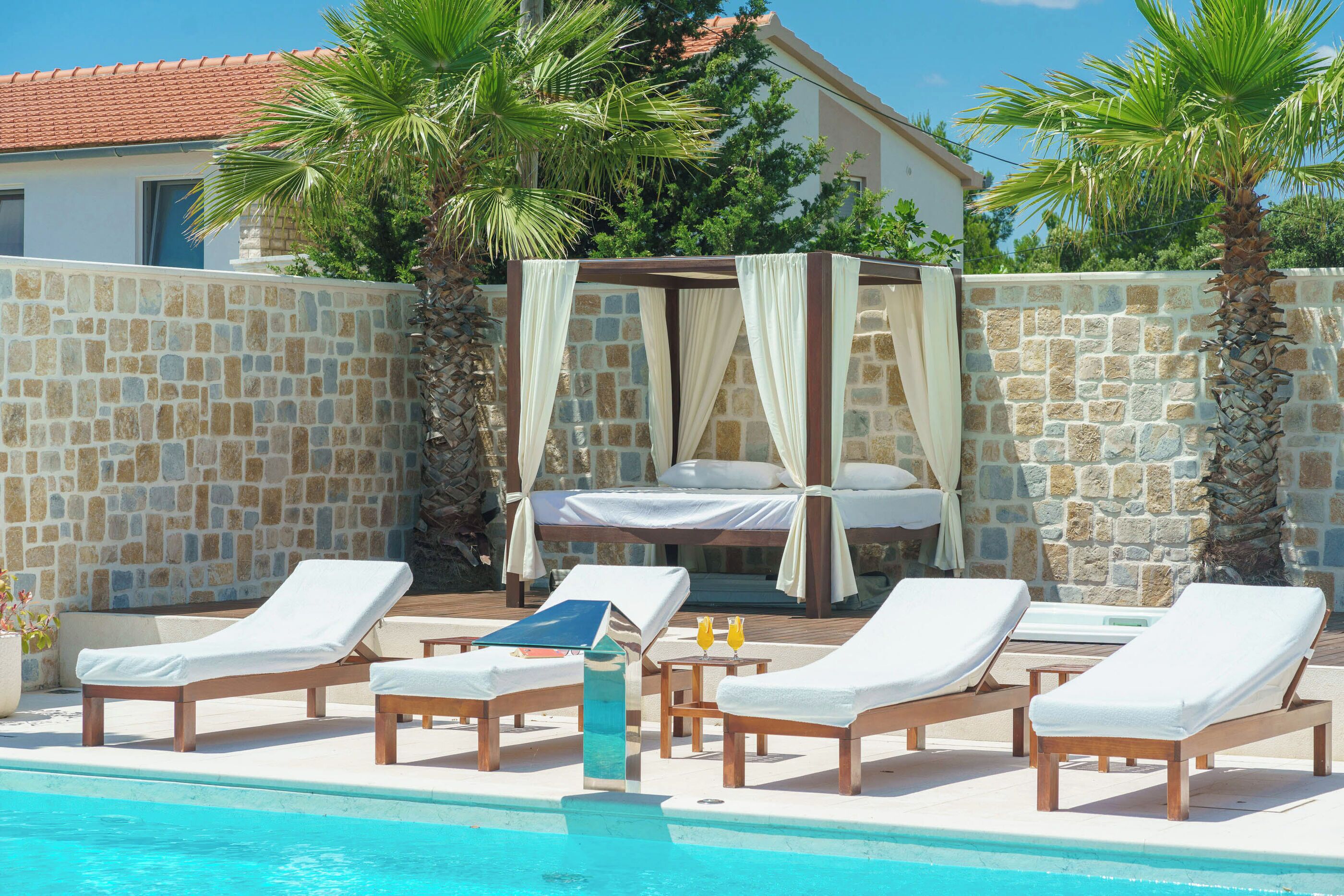 A poolside area with lounge chairs, a bed with a canopy, and a stone wall.