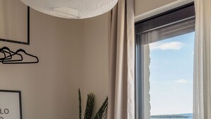 Une chambre à coucher comprenant un lit, une table de chevet, un luminaire suspendu, une fenêtre avec des rideaux et une vue sur la mer.