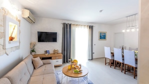 A modern living room with a sofa, dining table, and chairs. There is a flat-screen TV mounted on the wall, a small table with a plant, and a window with curtains. - Villa Patria (Rogoznica)
