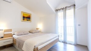 A bedroom with a wooden bed, a nightstand, a wall-mounted air conditioner, a painting, and a window with sheer curtains. - Villa Patria (Rogoznica)