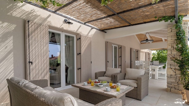Eine sonnendurchflutete Terrasse mit Korbmöbeln, einem gedeckten Frühstückstisch und einer Steinmauer mit Kletterpflanzen.