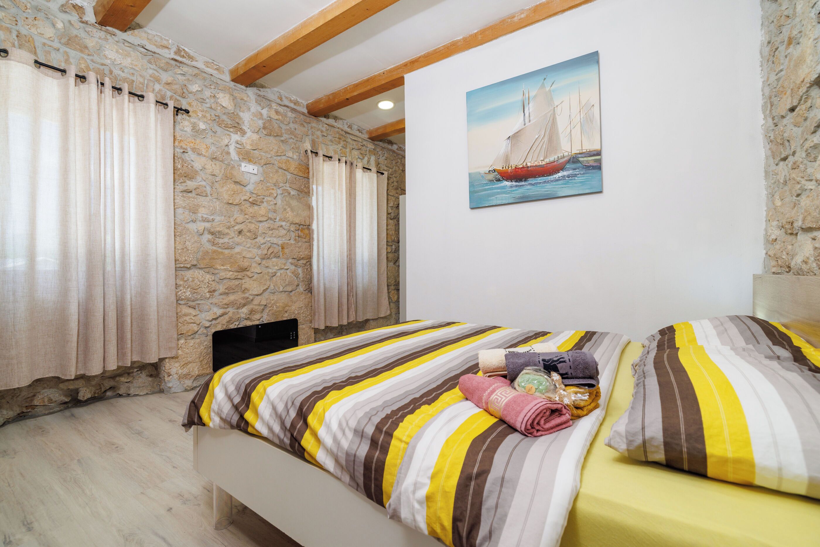 Een slaapkamer met een stenen muur, een schilderij van een zeilboot, een bed met gestreept beddengoed, en een bankje.