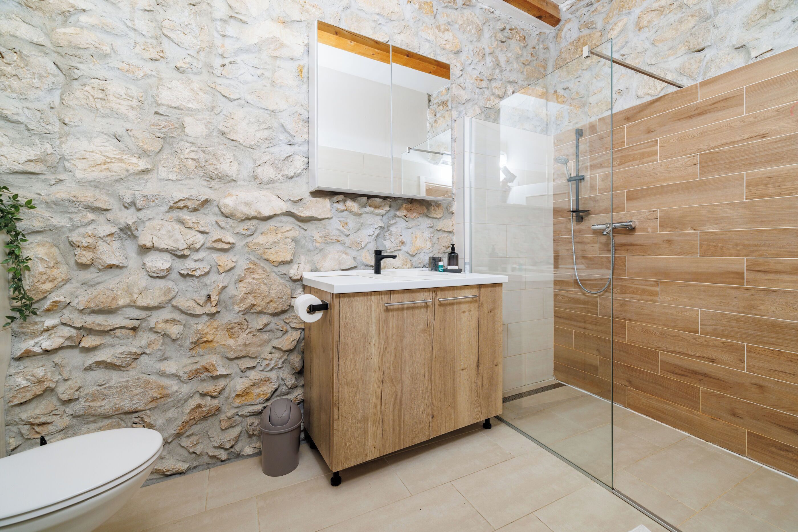 Une salle de bain avec un mur en pierre, des meubles en bois, une cabine de douche en verre et un lavabo blanc.