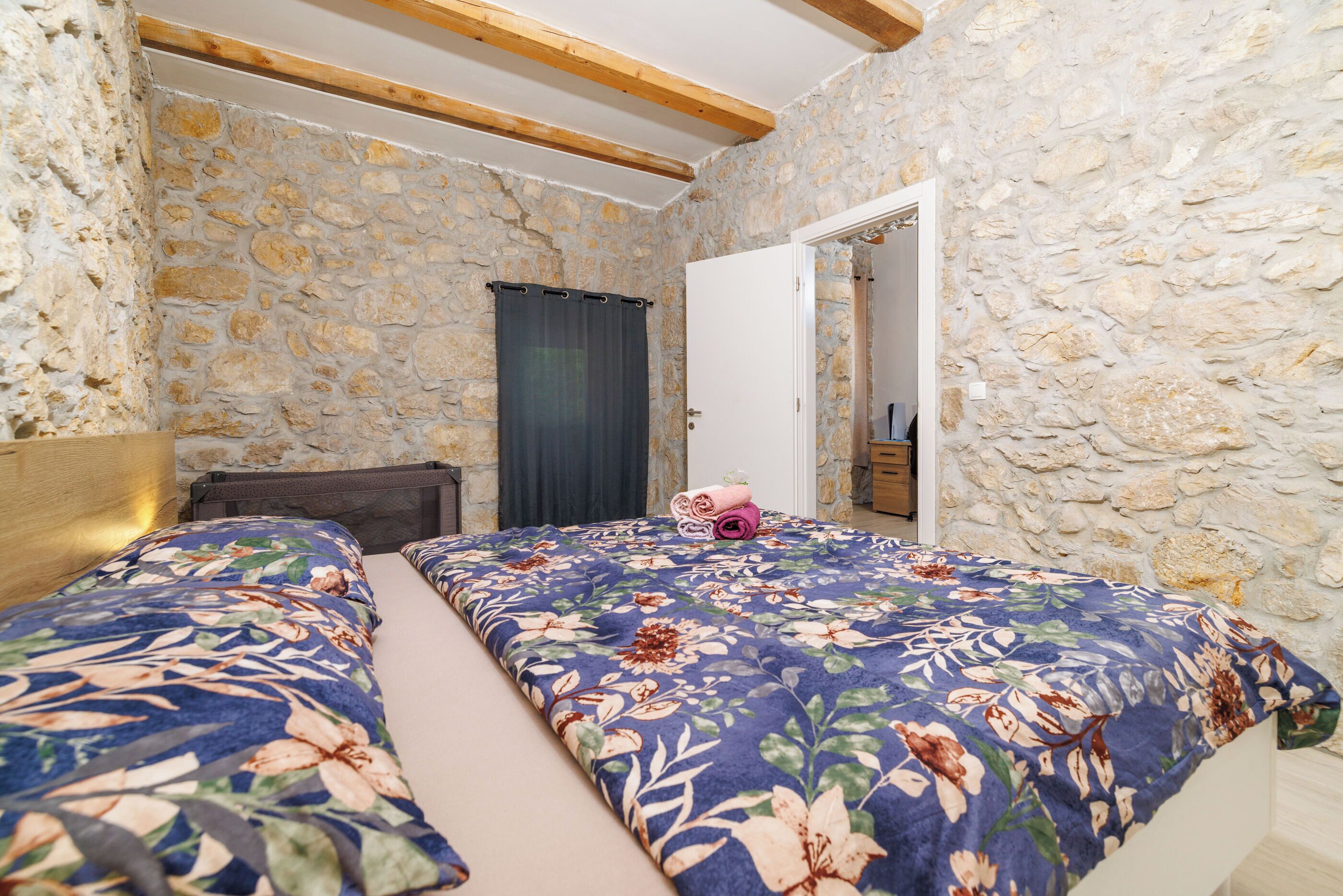 A bedroom with stone walls, wooden beams, two beds with floral bedding, a nightstand, and a door leading to another room.