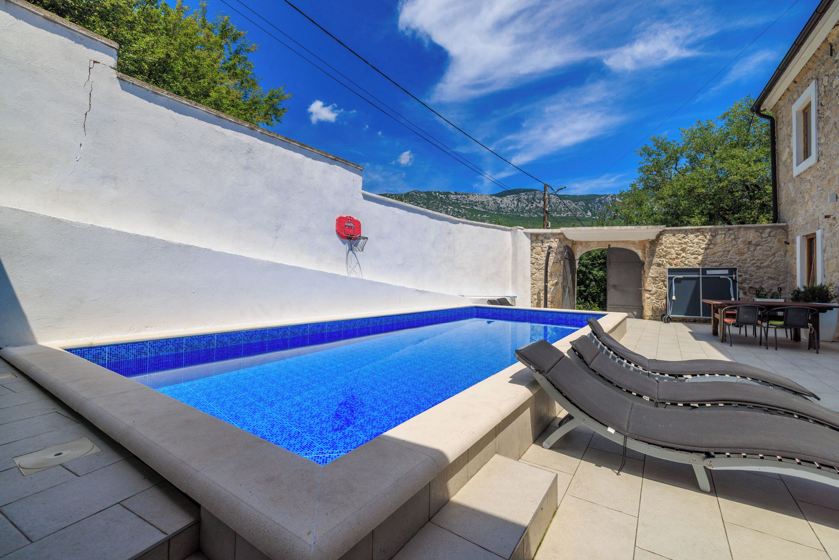 Una piscina con lettini, un canestro da basket rosso e un muro in pietra.