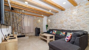 A living room with a stone wall, wooden ceiling beams, a grey sofa, a wooden coffee table, and a TV mounted on the wall. - Villa Stone Castle (Vinodol)