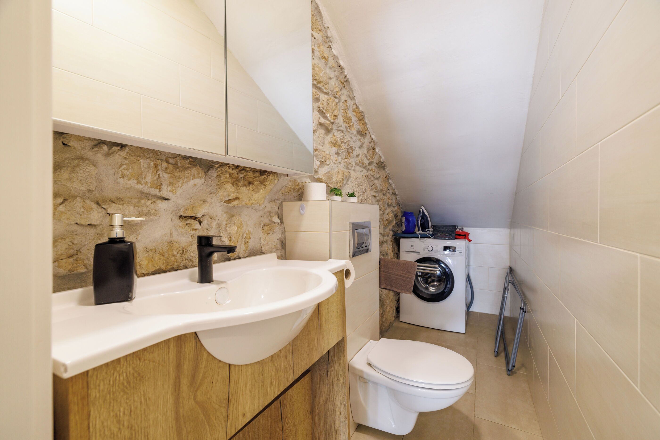 A compact bathroom with a sink, toilet, and washing machine, featuring stone wall accents and wooden cabinetry.