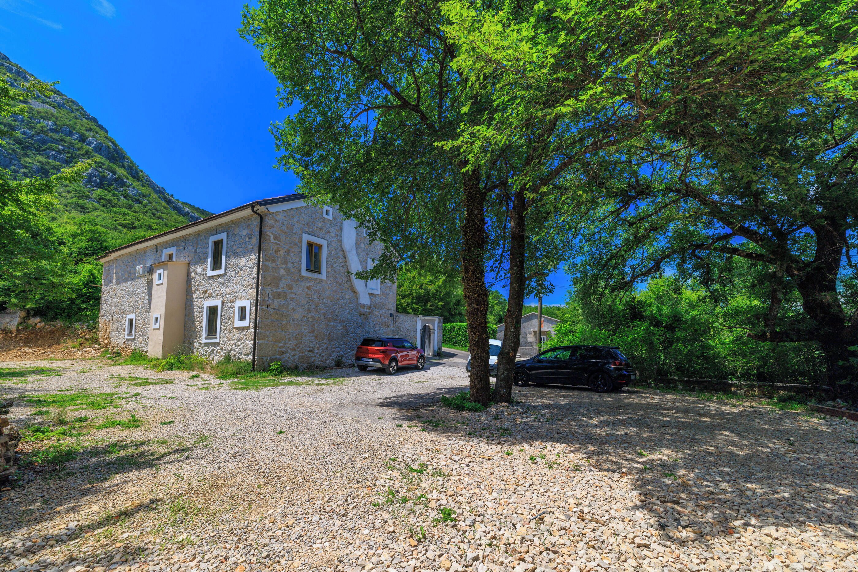 Un bâtiment en pierre avec une allée de gravier, des voitures garées et une chaîne de montagnes en arrière-plan.