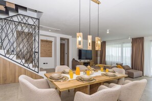 A modern dining area with a wooden table set for a meal, a staircase with a geometric railing, and a living area with a sofa and curtains. - Villa El Viento (Kraljevica)
