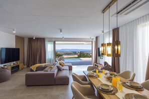Een moderne woonkamer met een grote bank, een eettafel gedekt voor het ontbijt, en uitzicht op een zwembad en bergen.