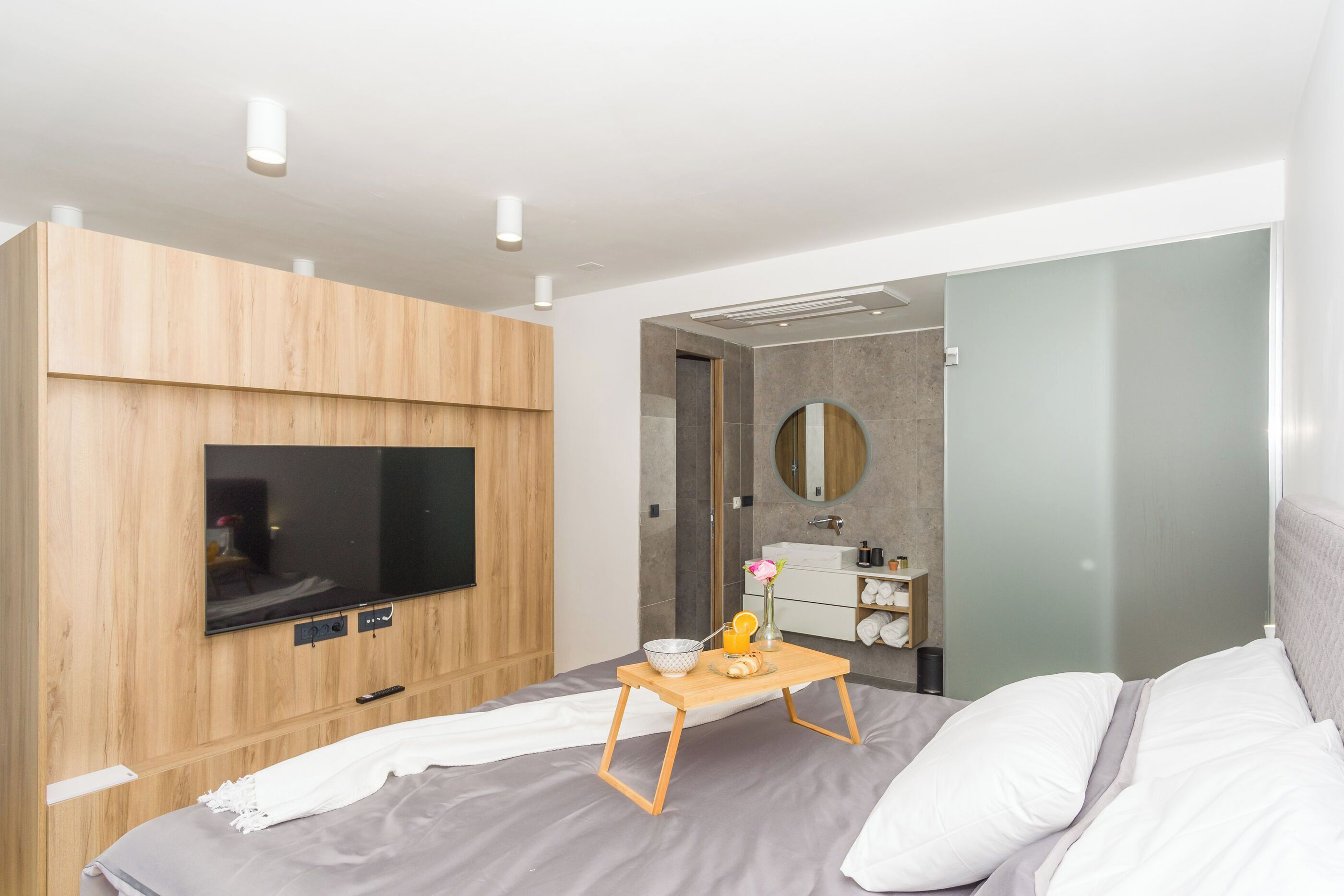 Une chambre moderne avec un lit, une armoire en bois, une télévision, une table de chevet avec un plateau, et une salle de bain visible à travers une porte ouverte.