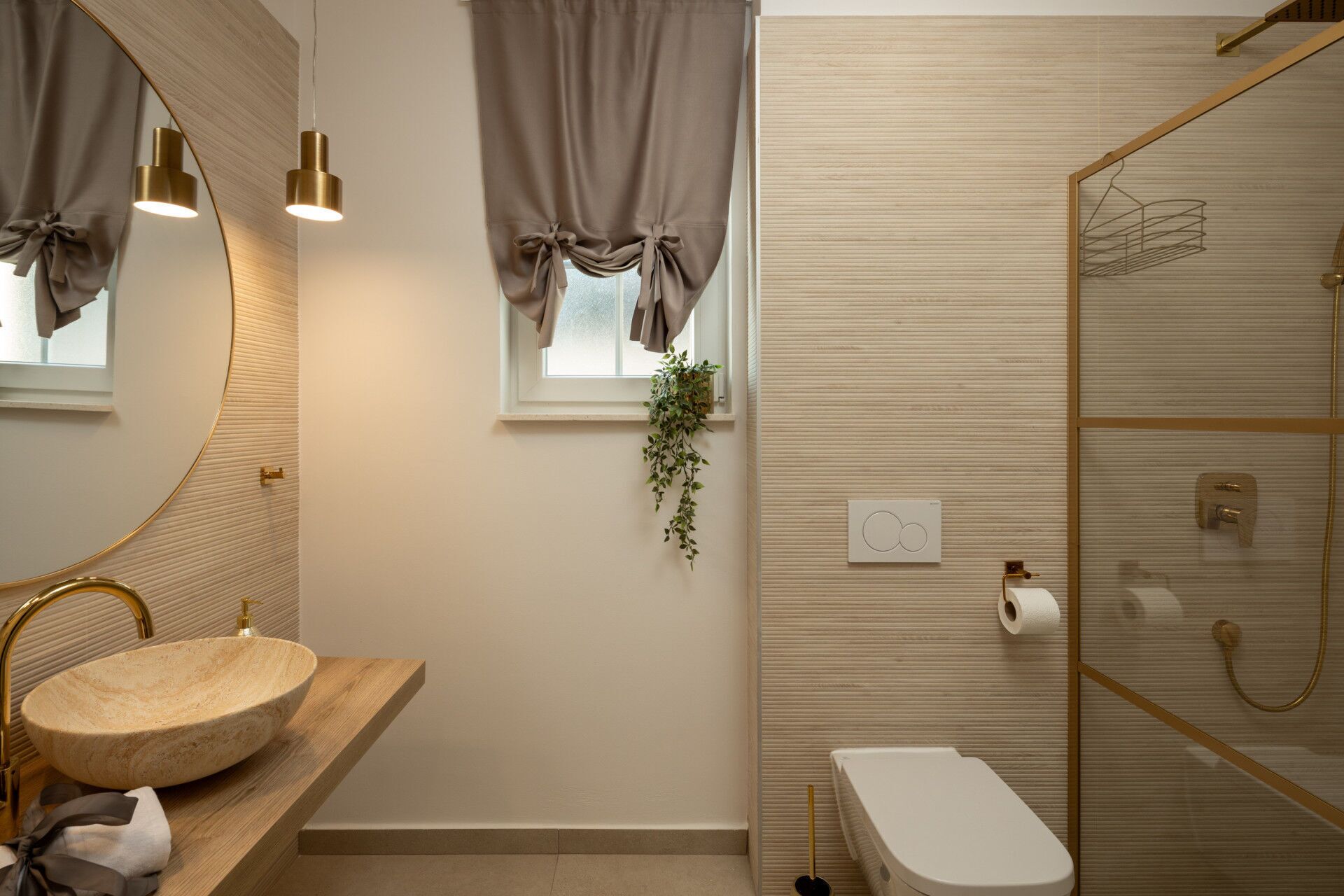 A modern bathroom with a round sink, gold fixtures, a shower with a glass door, and a window with curtains.