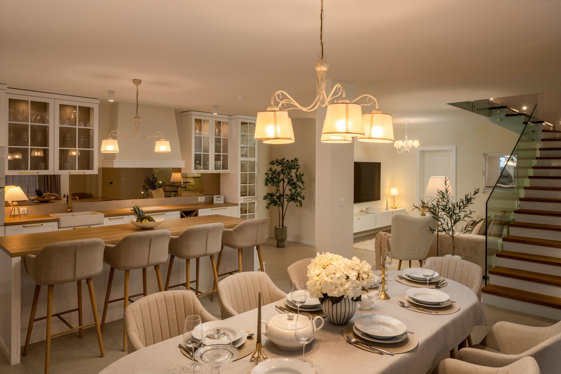 A modern dining area with a long table set for a meal, surrounded by comfortable chairs. A chandelier hangs above, and a staircase is visible in the background.