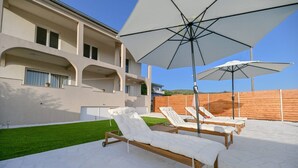 A modern outdoor lounge area with sun loungers and umbrellas, a well-maintained lawn, and a large building in the background. - Villa Benedicta (Pakostane)