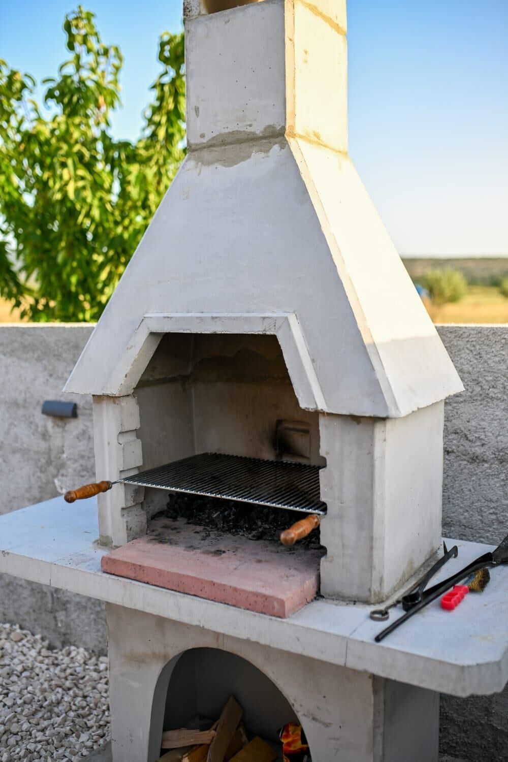 An outdoor brick oven with a metal grill, a chimney, and a small shelf with tools.