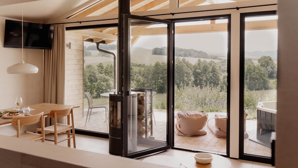 A modern interior with a large sliding glass door, a flat-screen TV, a dining table, and a cozy seating area with cushions. - Cabin Chalets House THREE (Geisa)