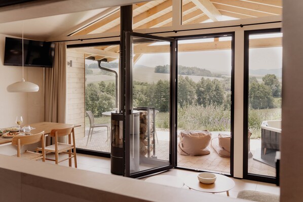 A modern interior with a large sliding glass door, a flat-screen TV, a dining table, and a cozy seating area with cushions. - Cabin Chalets House ONE (Geisa)