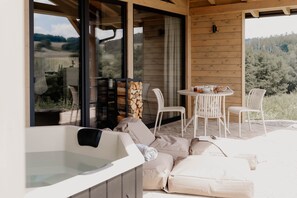 A wooden deck with a hot tub, white chairs, and a table with a view of the forest. - Cabin Chalets House ONE (Geisa)