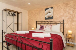 Una cama bien hecha con una colcha roja, estructura metálica y un perchero de pared con ganchos.