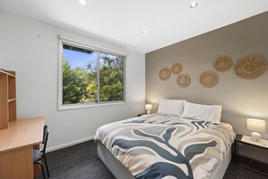 A bedroom with a bed, a desk, a chair, a window with a view of trees, and wall decor. - Point View Beach House (Lorne)