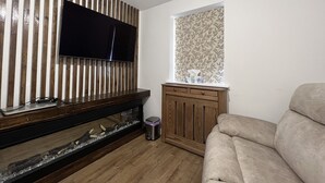 Una sala de estar con una televisión de pantalla plana montada en una pared de listones de madera, una ventana con estampado floral, un mueble de madera y un sofá color beige.