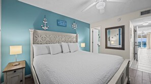 A bedroom with a bed, a nightstand, a ceiling fan, a wall-mounted ship's wheel, and a door leading to a balcony. - Ground level Gulf front condo, w/ pool, grill & gazebo ~ Sleeps 4 (Orange Beach)