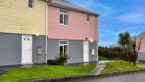 Een woonhuis met twee verdiepingen, een gevel in grijs en roze, witte deuren en een kleine voortuin met gras en een palmboom.