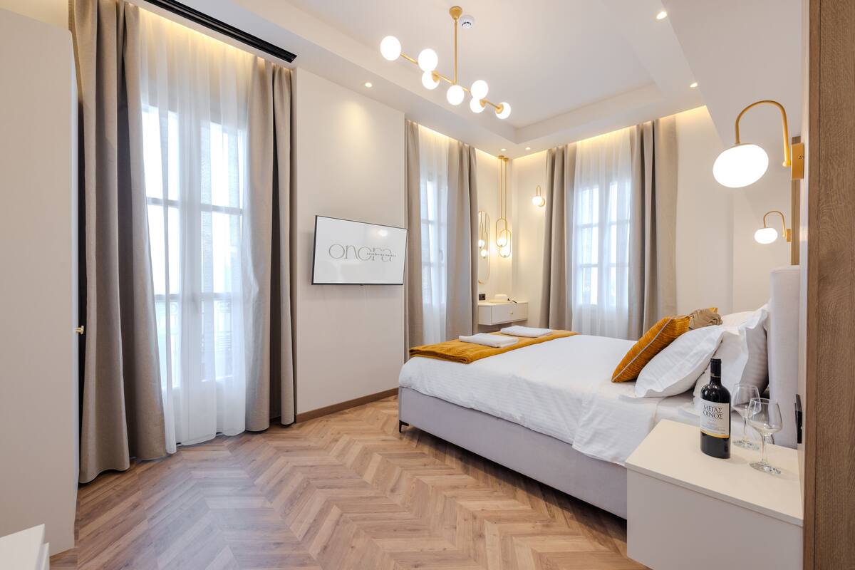 A modern hotel room with a large bed, a bedside table with a bottle of wine and glasses, and a wall-mounted TV.