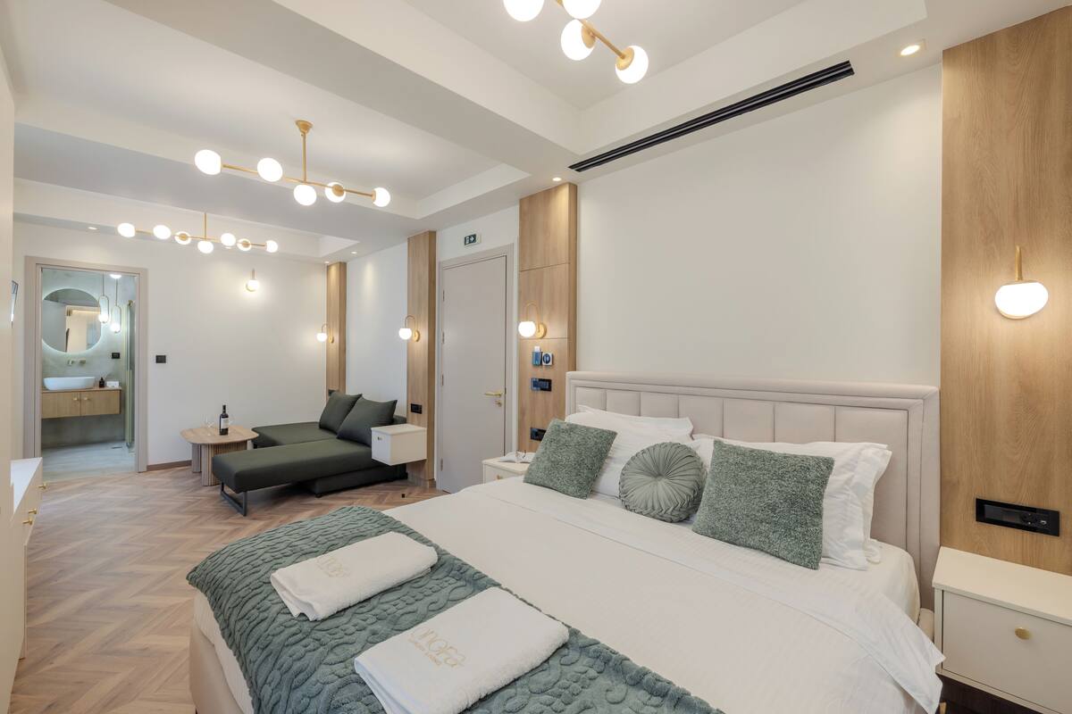 A modern hotel room with a large bed, a sofa, a vanity with a mirror, and a small table with a bottle.