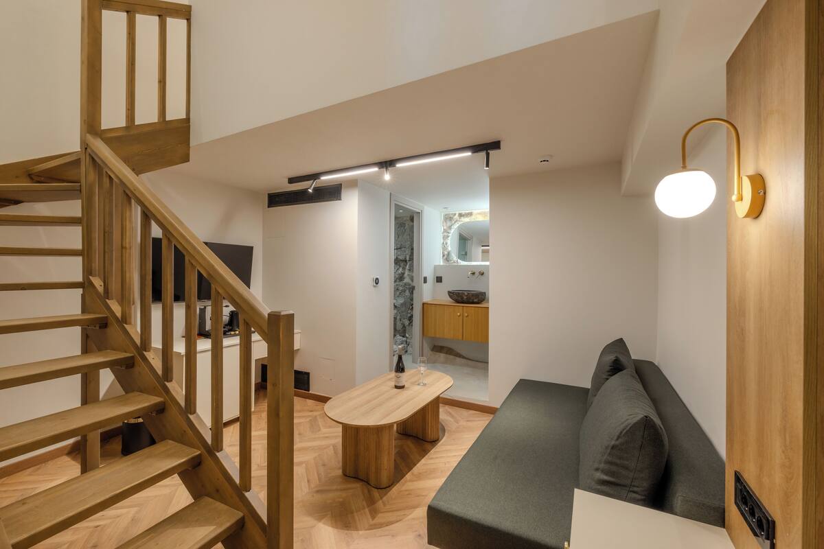 A modern interior with a wooden staircase, a dining area with a table and chairs, a sofa, and a kitchen area with a sink and countertop.