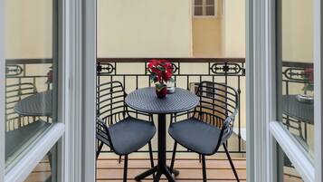 Varanda aberta com mesa e cadeiras, um vaso de planta e vista para um edifício com telhado em azulejos.