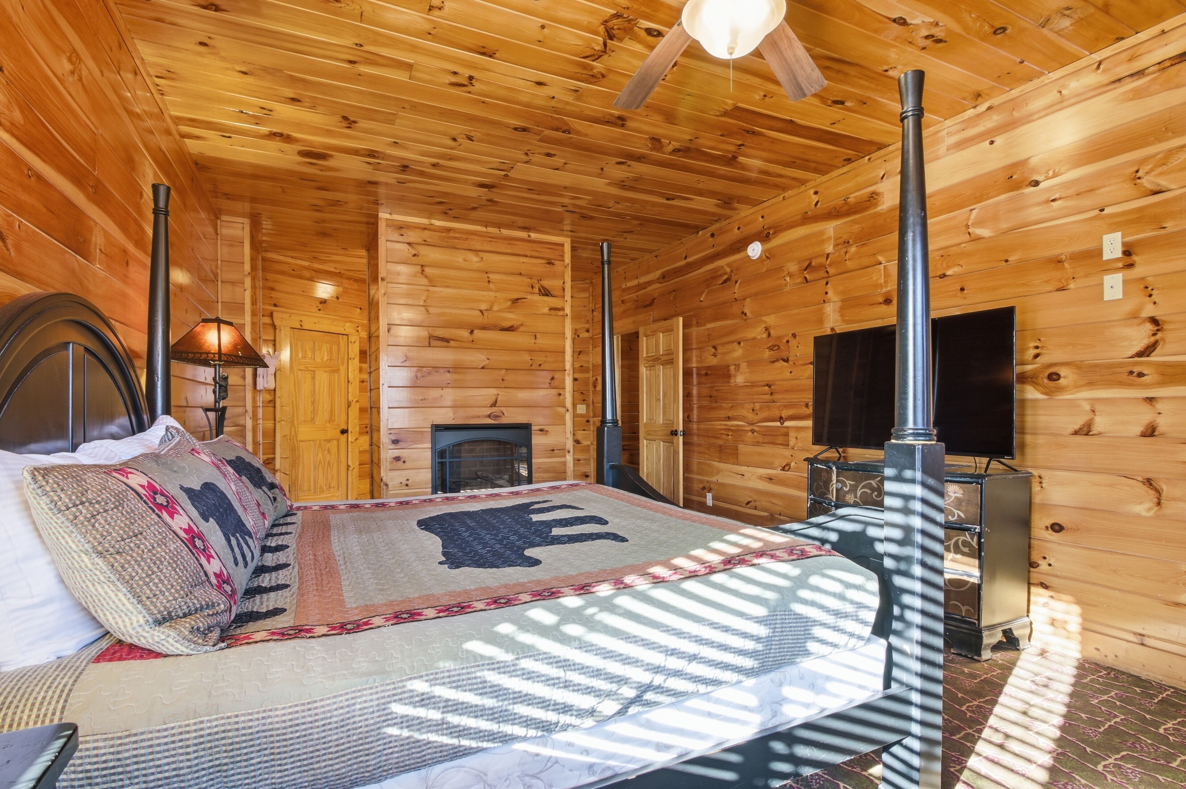 A log cabin bedroom with a bed, a fireplace, a television, and a ceiling fan.