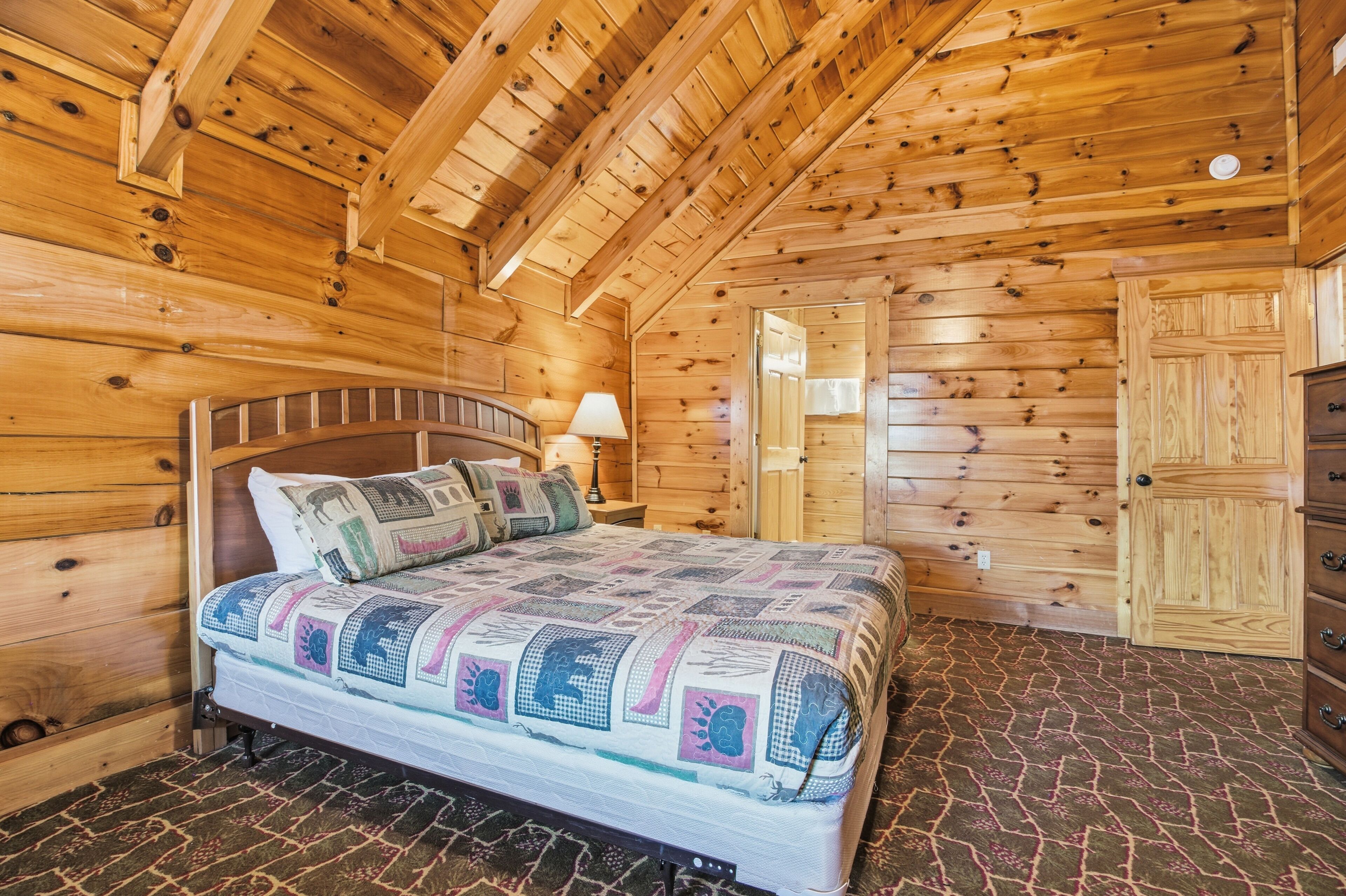 A wooden cabin bedroom with a bed, nightstand, lamp, dresser, and a door.