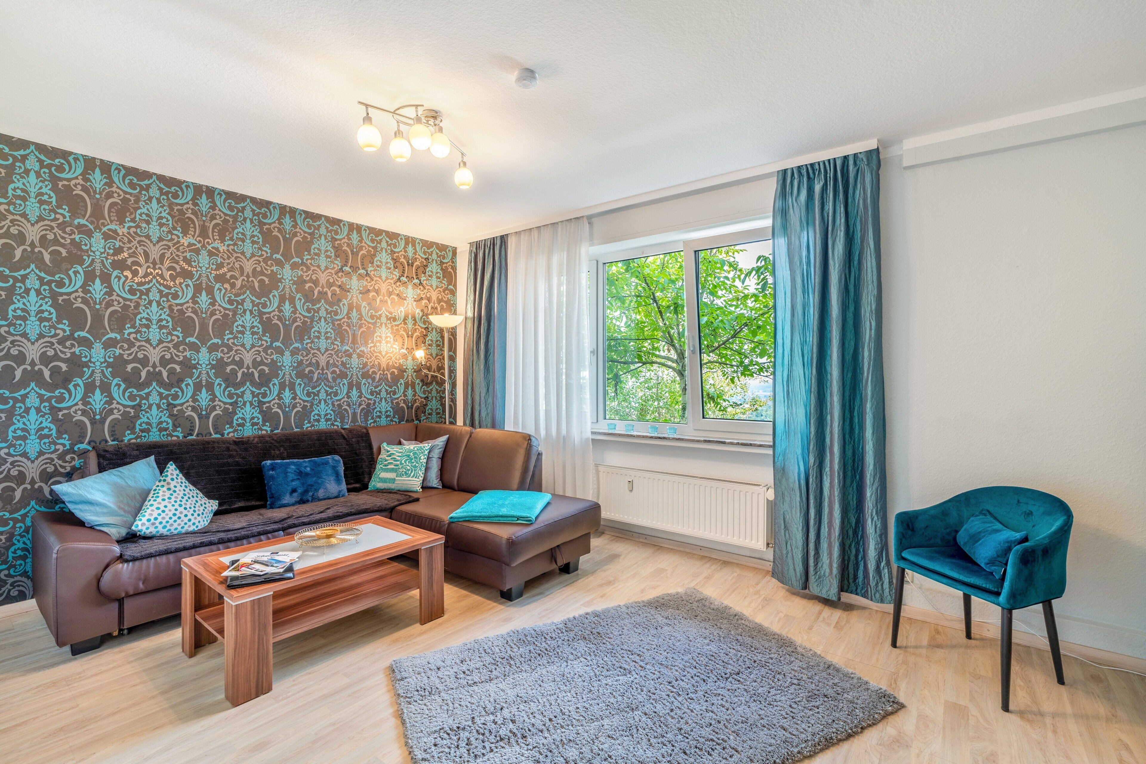 A living room with a brown sofa, a coffee table, a teal armchair, and a window with curtains.