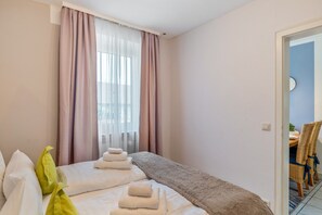 Una habitación de hotel con una cama, un ventanal con cortinas, una puerta que da a un comedor y un reloj de pared.