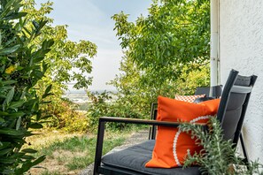 Uma cadeira com almofada, posicionada perto de uma parede branca, com vista para vegetação e uma paisagem distante.